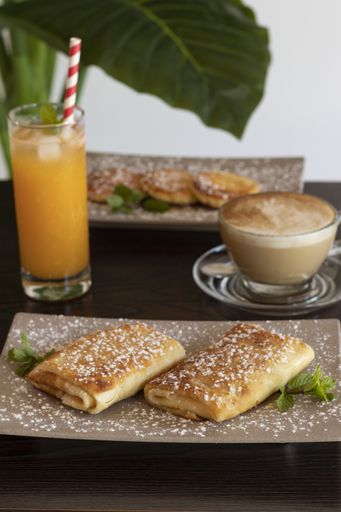 A vertical shot of a breakfast table with syrniki, pancake rolls, orange juice and coffee