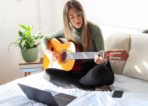 An attractive young woman sitting on the bed and playing acoustic guitar with a laptop in front of her