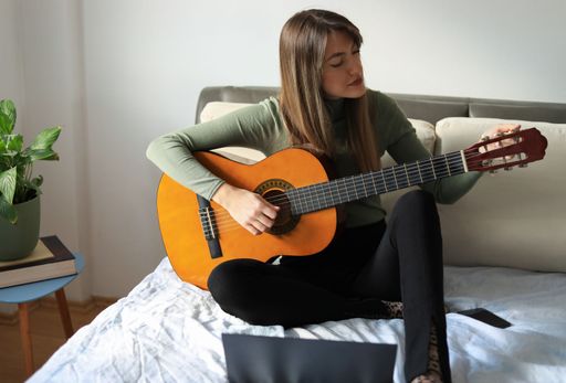 An attractive young woman sitting on the bed and playing acoustic guitar with a laptop in front of her
