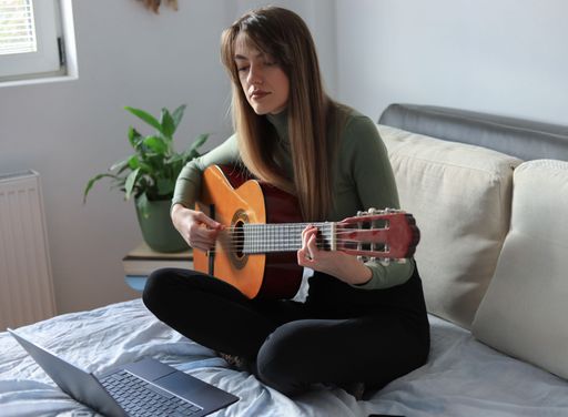 An attractive young woman sitting on the bed and playing acoustic guitar with a laptop in front of her
