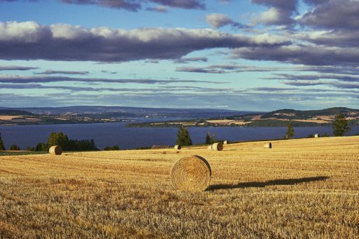 The floating clouds above a hay field with straw bales and Lake Mjosa in the distance in Toten, Norway