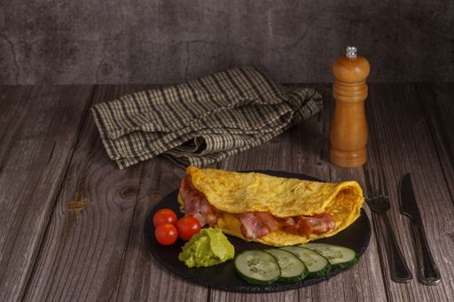 bacon omelet on a round black slate with tomato, guacamole and cucumber garnish with pepper shaker and cutlery on a wooden table