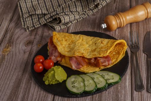 bacon omelet on a round black slate with tomato, guacamole and cucumber garnish with pepper shaker and cutlery on a wooden table