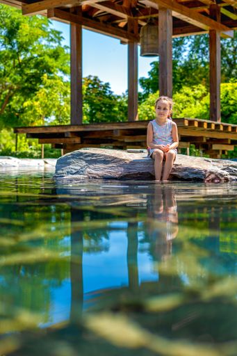 A vertical shot of a pretty little Hispanic girl sitting on a stone by the water
