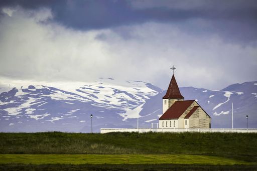 The Vik i Myrdal Church by lush green fields in Iceland with snowcap mountains in the background