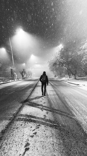 A vertical grayscale shot of a lone male walking on a snowy road under street lights