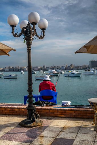 A back view of male fishing in lake surrounded by boats and buildings in Alexandria