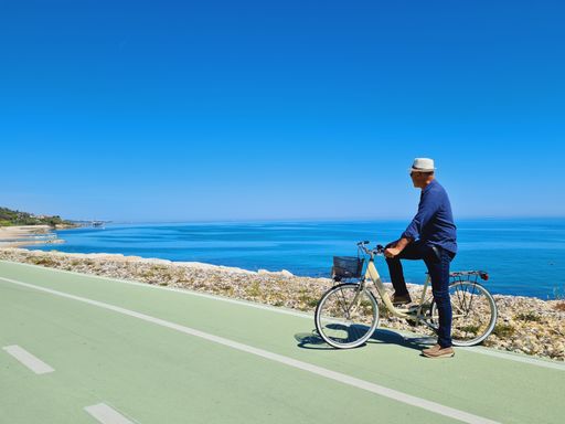 A man on a bike, standing on the road next to the rocky Trabocchi coast, with a blue calm sea in the background, on a sunny day in Italy
