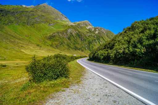 A landscape of a road surrounded by hills covered in greenery under a blue sky and sunlight in Tyrol