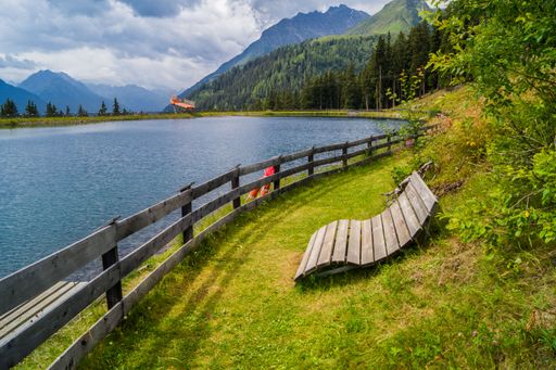 A wooden bench on the lakeside surrounded by lush greenery. Silvretta High Alpine Road, Switzerland.
