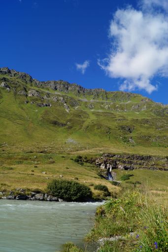 A vertical shot of a small waterfall in a green valley with a cloudy blue sky in the background