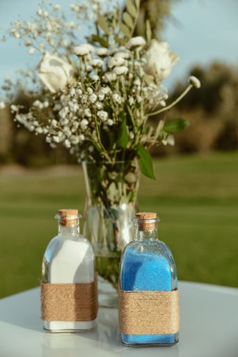 A vertical shot of glass bottles with colored sand on a table with a vase and a floral bouquet