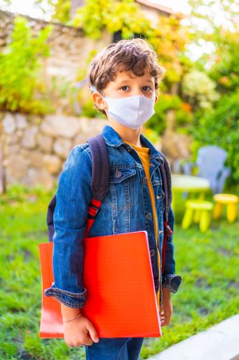 A shallow focus of a kid wearing a facemask and holding his school belongings under the sunlight