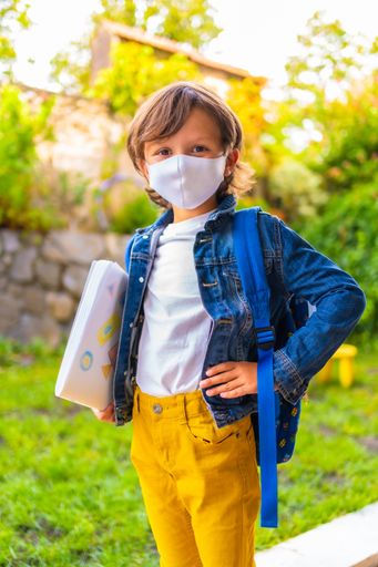A shallow focus of a kid wearing a facemask and holding his school belongings under the sunlight