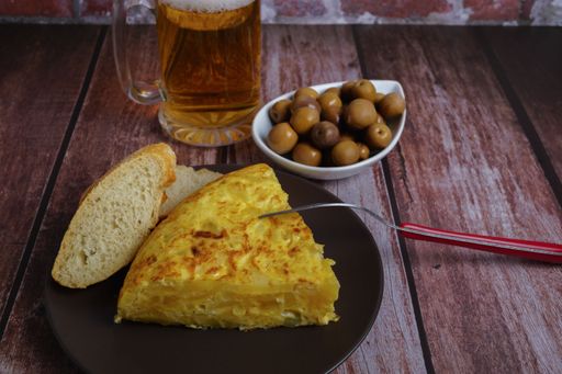 typical spanish potato omelette with bread, olives, cutlery and beer mug on a wooden table with a brick wall in the background.
