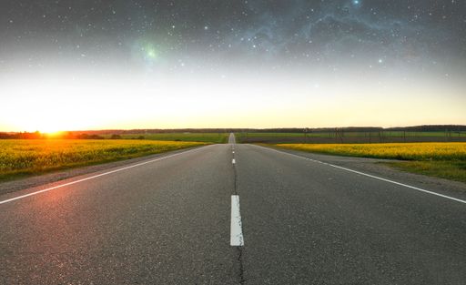An empty highway between green fields at sunset under a starry sky