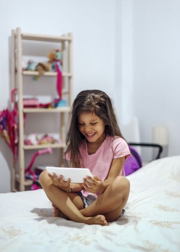 A vertical shot of a Hispanic female child  having fun playing on a tablet in her room