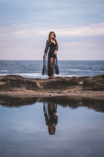 A young brunette Caucasian female in a long black transparent dress on some rocks near the sea on a summer afternoon- reflecting in the sea
