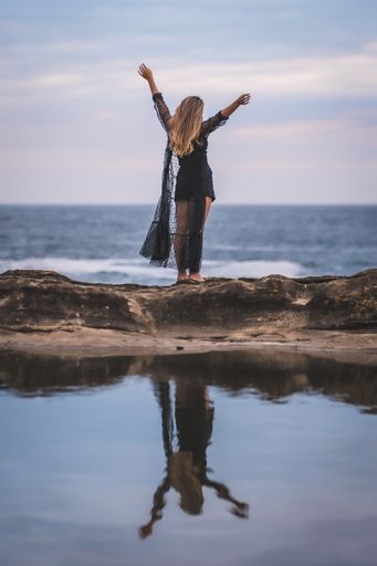 A young brunette Caucasian female in a long black transparent dress on some rocks near the sea on a summer afternoon- reflecting in the sea