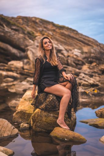 A young brunette Caucasian female in a long black transparent dress on some rocks near the sea on a summer afternoon- reflecting in the sea