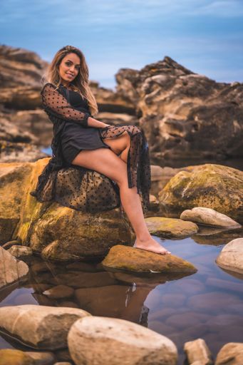 A young brunette Caucasian female in a long black transparent dress on some rocks near the sea on a summer afternoon- reflecting in the sea