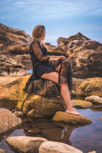 A young brunette Caucasian female in a long black transparent dress on some rocks near the sea on a summer afternoon- reflecting in the sea
