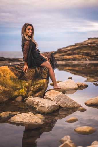 A young brunette Caucasian female in a long black transparent dress on some rocks near the sea on a summer afternoon- reflecting in the sea