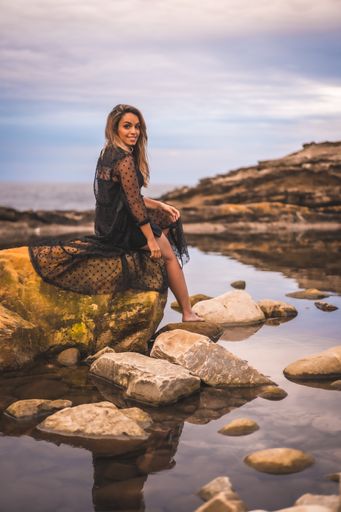 A young brunette Caucasian female in a long black transparent dress on some rocks near the sea on a summer afternoon- reflecting in the sea