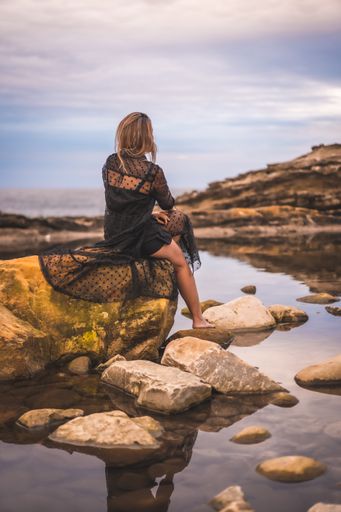 A young brunette Caucasian female in a long black transparent dress on some rocks near the sea on a summer afternoon- reflecting in the sea