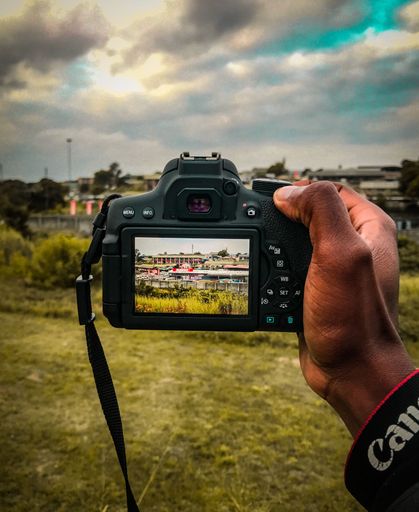 A vertical shot of a hand holding a Canon camera, photographs a rural landscape under cloudy sky