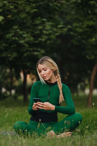 A vertical shot of a young female with a phone in her hands typing and smiling in the green park