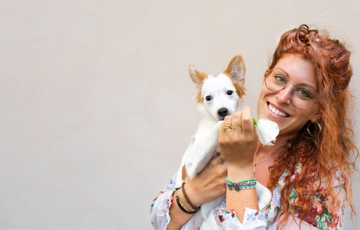 A young woman with long, red hair and glasses is cradling a small, white and brown dog in her arms.
