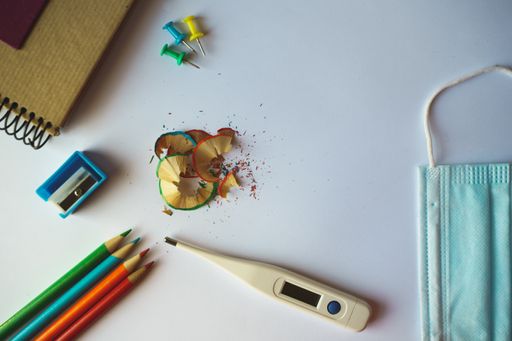 A high angle shot of school supplies, a medical mask, and a thermometer on a white surface - back to school concept in the new normal after Covid-19