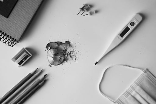 A grayscale shot of school supplies, a medical mask, and a thermometer on a white surface - back to school concept in the new normal after Covid-19