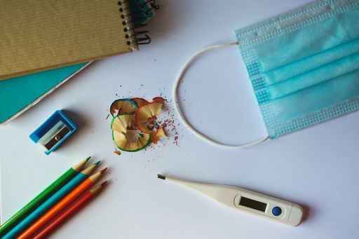 A high angle shot of school supplies, a medical mask, and a thermometer on a white surface - back to school concept in the new normal after Covid-19