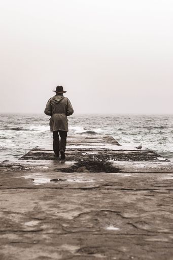 A grayscale back shot of a coat man with hat standing by a rough sea under light sky