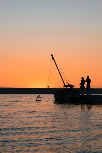 A silhouette view of a couple standing at the shore by a docked boat against dusk sky