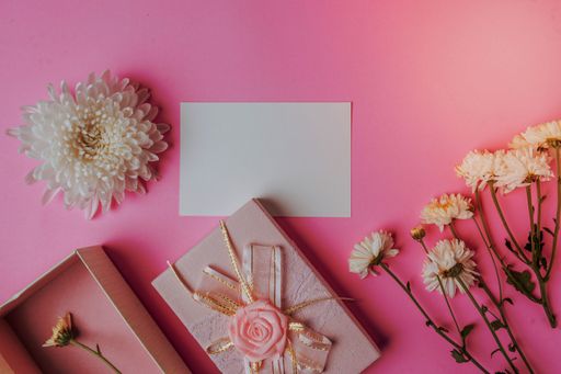 A pink background with a piece of paper sitting next to flowers and a gift box, with copy space
