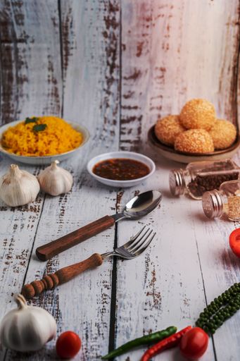 A spoon, fork chili sauce with spices, garlic heads, and cooked rice on a white wooden floor