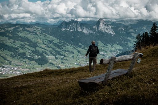 A hiker standing by a wooden bench on a slope facing the scenic view of the mountains