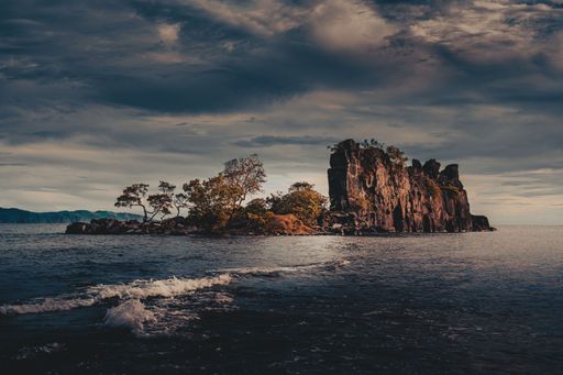 A small island seen from the shore with rock formation and few trees under dark cloudy sky