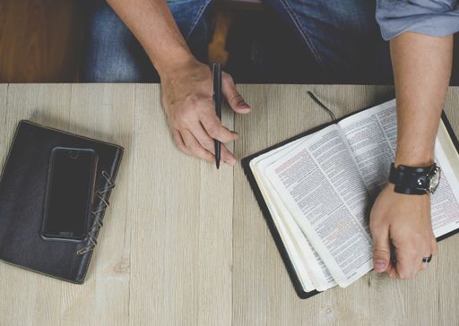 An overhead shot of a male reading the bible and holding a pen near his notebook and smartphone