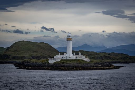 A view of the Lismore Lighthouse on the edge of a green coast before the hills under the cloudy sky