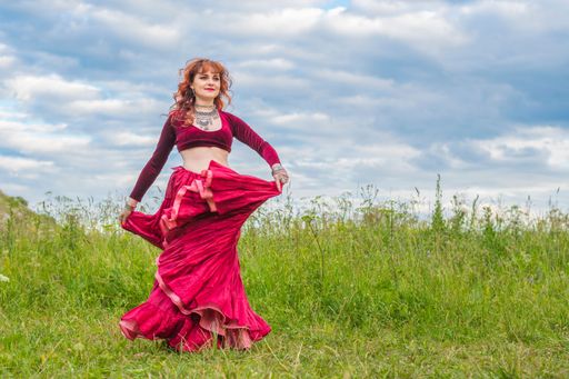 Young red-haired girl in red ball gown dances incendiary dance on green meadow against blue evening summer cloudy sky.