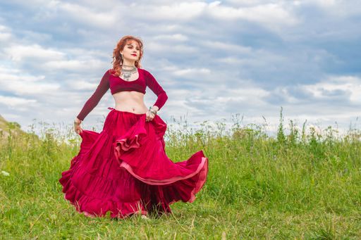 Young red-haired girl in red ball gown dances incendiary dance on green meadow against blue evening summer cloudy sky.