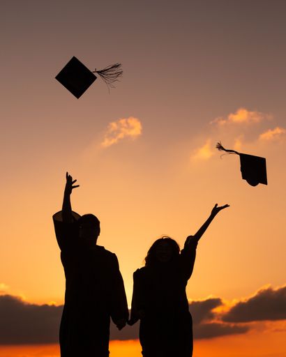 The silhouettes of two students throwing their mortarboards in the air at the graduation on the background of an orange sunset