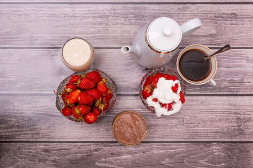 On wooden table are white teapot, two crystal vases with ripe red strawberries, two glasses filled with cocktails, and cup of tea or coffee