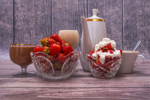 On wooden table are white teapot, two crystal vases with ripe red strawberries, two glasses filled with cocktails, and cup of tea or coffee.