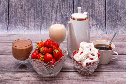 On wooden table are white teapot, two crystal vases with ripe red strawberries, two glasses filled with cocktails, and cup of tea or coffee.