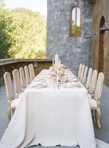 A long wedding reception table with white cloth cover with trees in the background outdoors
